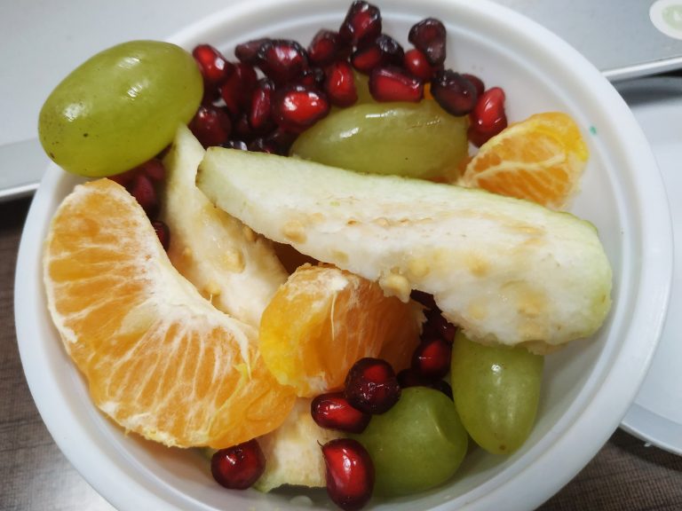 A bowl of mixed fresh fruit including green grapes, pomegranate seeds, and orange segments on a table.