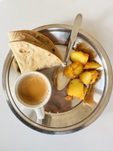 A stainless steel plate with a portion of flatbread, cooked potatoes, and a spoon, alongside a cup of tea on a white surface.