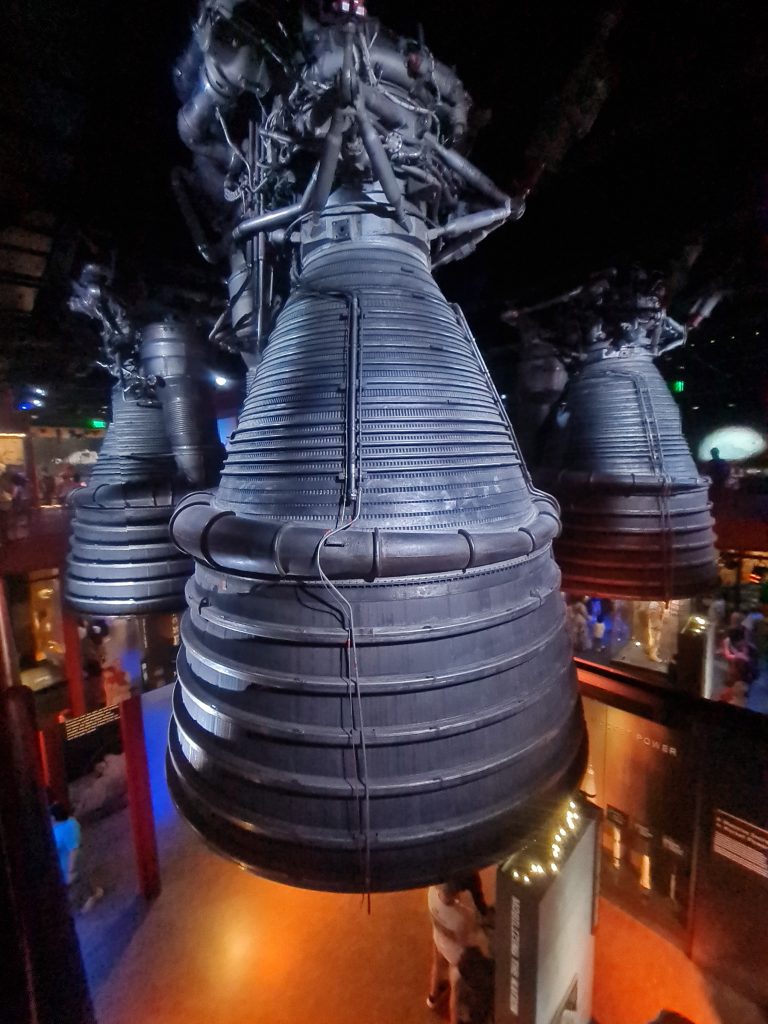 A large rocket engine exhibit suspended from the ceiling with intricate details and several people observing the exhibit from beneath, illuminated by dim lighting.