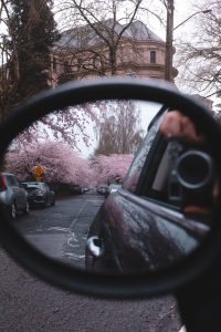 Looking back through a side mirror of a car reveals a row of cherry blossom trees in bloom on a neighborhood street lined with cars. The camera taking the photo is barely in view in the side mirror. 
