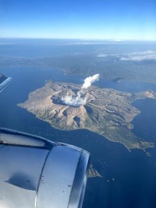 An aerial view from an airplane window showcasing Taal Volcano in Batangas, Philippines with a plume of smoke rising from the crater, surrounded by a body of water, with land and clouds in the distance.