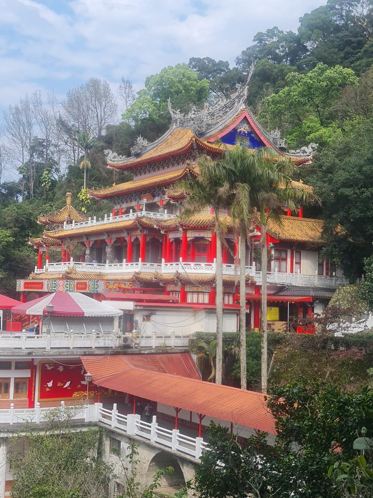 This photo captures a colorful multi-tiered traditional Chinese temple nestled on a hillside, adorned with intricate architectural details and surrounded by lush greenery under a clear sky.