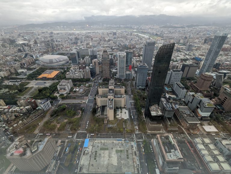 Taipei city landscape from the 88th floor. Showing many buildings and the Taipei stadium, with mountains in the background behind some cloud cover.