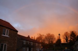A faint rainbow arcs over a quaint village street in Yorkshire, UK, with traditional stone buildings standing beneath an orange-tinted sky at sunset. In the foreground, there is a street lamp.