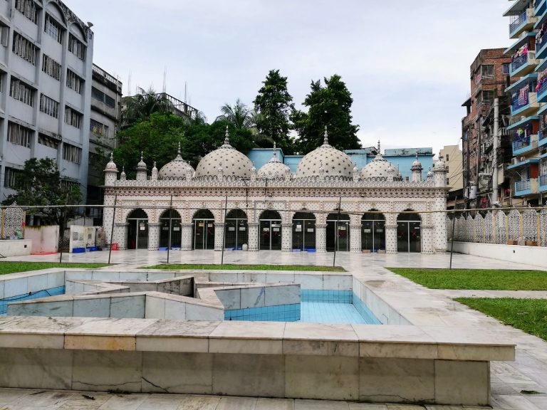 A mosque with multiple domes and minarets featuring intricate designs, in an urban environment with modern buildings in the background. Tara Masjid (Star Mosque) in Dhaka