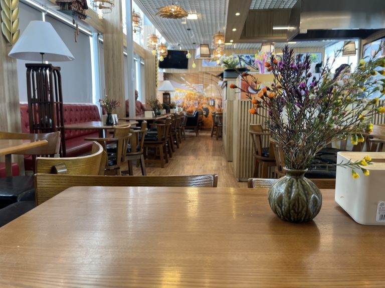 Interior of a restaurant with wooden tables and chairs, red upholstered booths, decorative lighting, and a vase with colorful flowers on a foreground table.