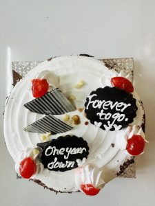 A round white anniversary cake with the inscriptions 'One year down' and 'Forever to go' on chocolate plaques, adorned with whipped cream dollops and red cherries, presented on a silver cake board.