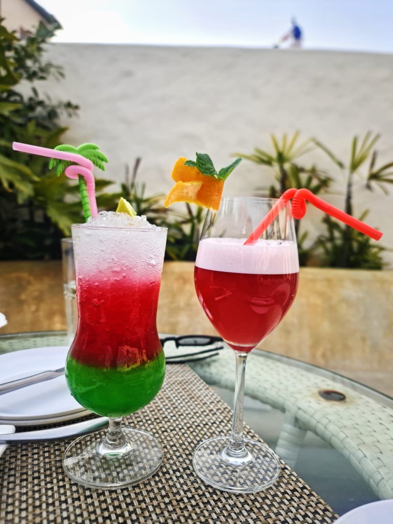 Two colorful cocktails on a table with a straw mat, one in a hurricane glass with a layered green, yellow, and red gradient and a pink straw, and one in a wine glass with a red beverage and orange slice garnish, both adorned with mint leaves.