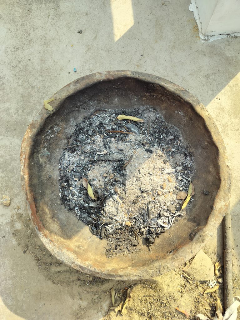 Ashes in a rustic hand thrown clay circular fire pit it sits outdoors on the dry dusty concrete floor . Three dried leaves have fallen on to the ash.