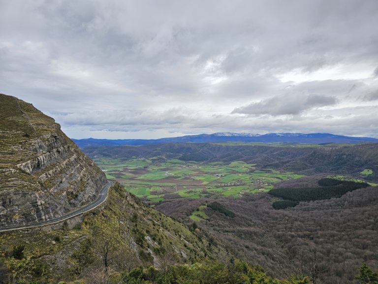 A panoramic view of a mountainous landscape from the BU-556 road on the Ordu?a mountain pass, with a winding road running alongside the slope on the left, overlooking green valleys with fields and scattered trees, with a backdrop of distant snow-capped mountains under a cloudy sky.