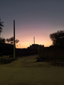 A sunset view over a rural street setting with silhouettes of trees, utility poles, wire cables, and rustic buildings against a gradient sky of deep blues and warm oranges.