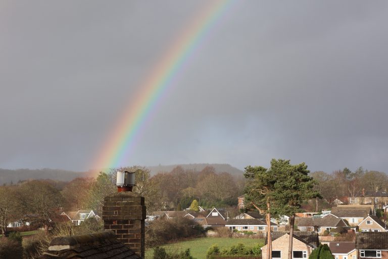 A rainbow over a grey sky. There are houses in the foreground.