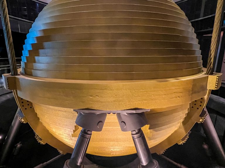 A close-up view of a wind damper in Taipei 101 which is a large metallic pendulum-like structure with concentric rings, suspended above a platform, supported by three symmetrically placed cylindrical dampers, with ambient lighting casting soft shadows and highlights on the surface.