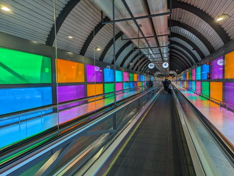 A moving walkway with vibrant, multicolored glass panels on the walls and ceiling, and passengers with luggage using the walkway.