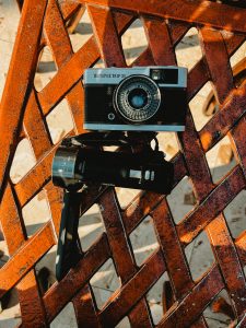 An old analog film camera resting on a rusted metal surface with geometric cut-out patterns, illuminated by warm sunlight.