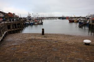 A harbour with multiple fishing boats. The water is calm. There is a stone gangway in the foreground.