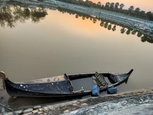 A dilapidated wooden boat partially submerged near the water's edge at sunset, with calm waters reflecting palm trees and the sky's orange hues.