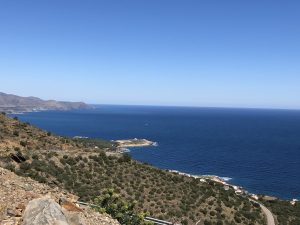  A panoramic view of a the Catalan coastal landscape with a clear blue sea, a curving shoreline, patches of greenery, and mountains in the distance under a bright blue sky.
