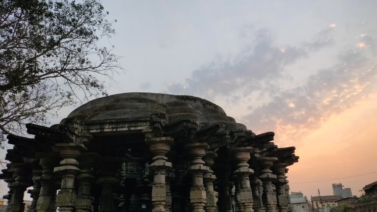 An ancient stone temple in Swargmandap Khidrapur, Maharashtra with multiple columns supporting a large dome, silhouetted against a sky with soft shades of orange and blue during sunset.