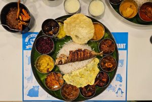  A large, Kerala-style non-veg thali meal served on a plate, with various dishes in small bowls, including rice, fried fish, papadum, and curries, on a table.
