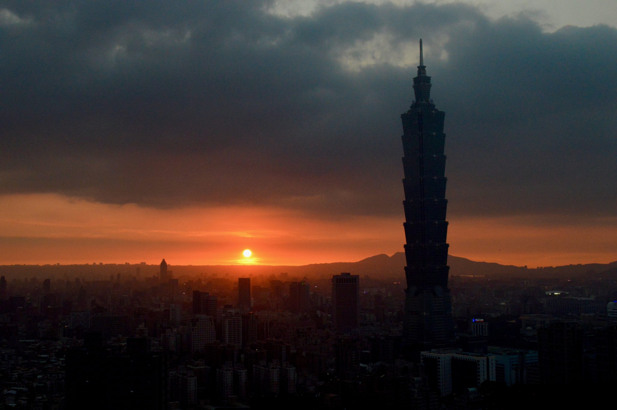 A wide city panorama of Taipei featuring the Taipei 101 skyscraper against a setting sun.