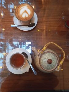 A cup of cappuccino with leaf pattern latte art on a saucer with a spoon and a cup of tea with a tea bag on the side, accompanied by a ceramic teapot with a rattan handle on a dark wooden table. Reflections and lights can be seen on the surface of the table.
