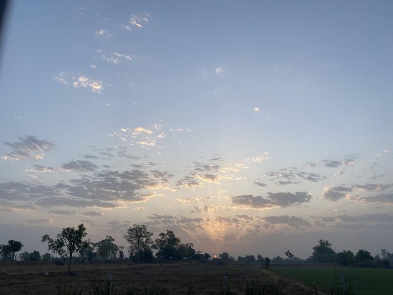 Rays of sunlight bursting through clouds at sunrise over a rural landscape with sparse trees and open fields.