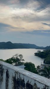 A scenic view of a lake surrounded by mountains during twilight, with a colorful sky above and the balcony edge of a building in the foreground.