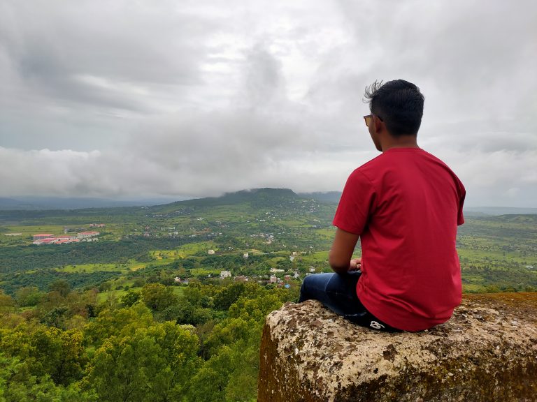 A boy captivated by nature’s beauty, seated on a rock, with a backdrop of a cloudy sky revealing a picturesque village and fort.