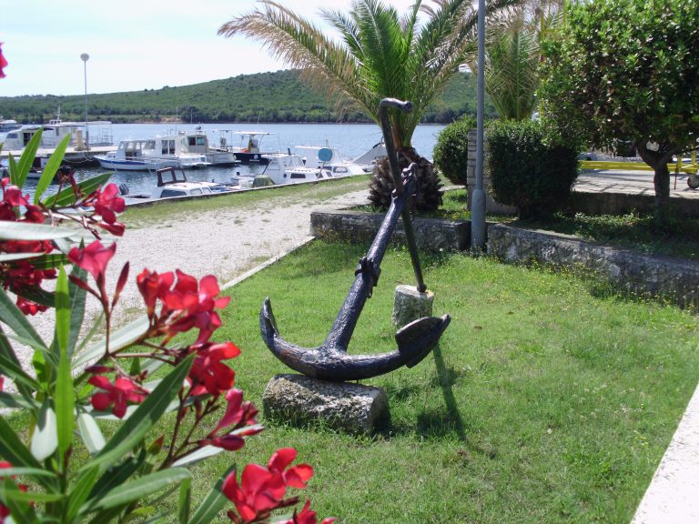 A large anchor displayed near a marina on grass near a walkway with oleander flowers in the foreground, palm trees and moored boats in a calm harbor in the background.