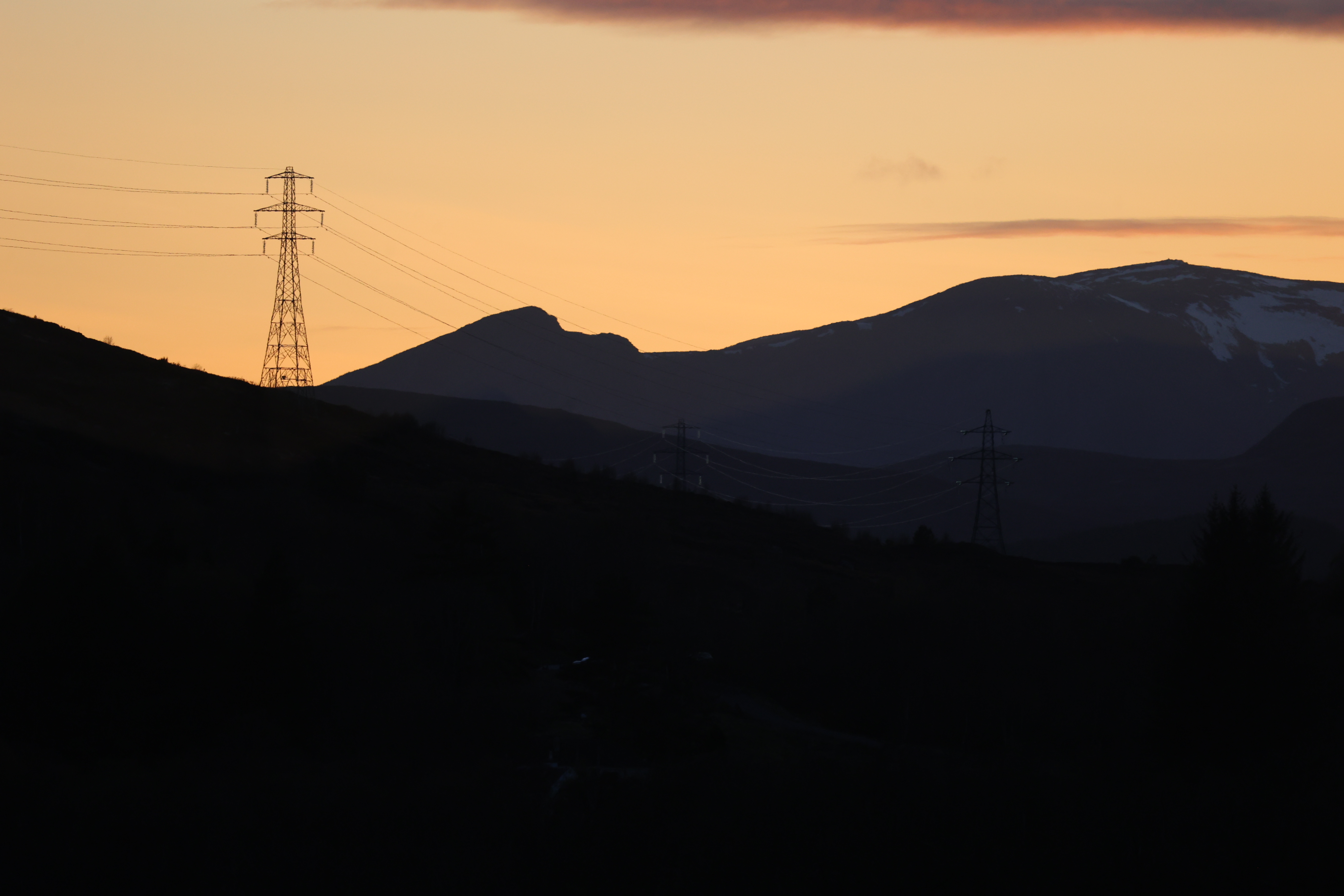 Electricity pylon in silhouette on a hill against a mountainous sunset sky, Scottish Highlands