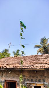 A weathered flag, mostly green with some yellow, fluttering at the top of a tall pole, with a backdrop of clear blue sky. The pole is in front of a traditional building with a red tiled roof and stone walls, with some green foliage and palm trees visible in the scene. (Gudi padwa festival) 