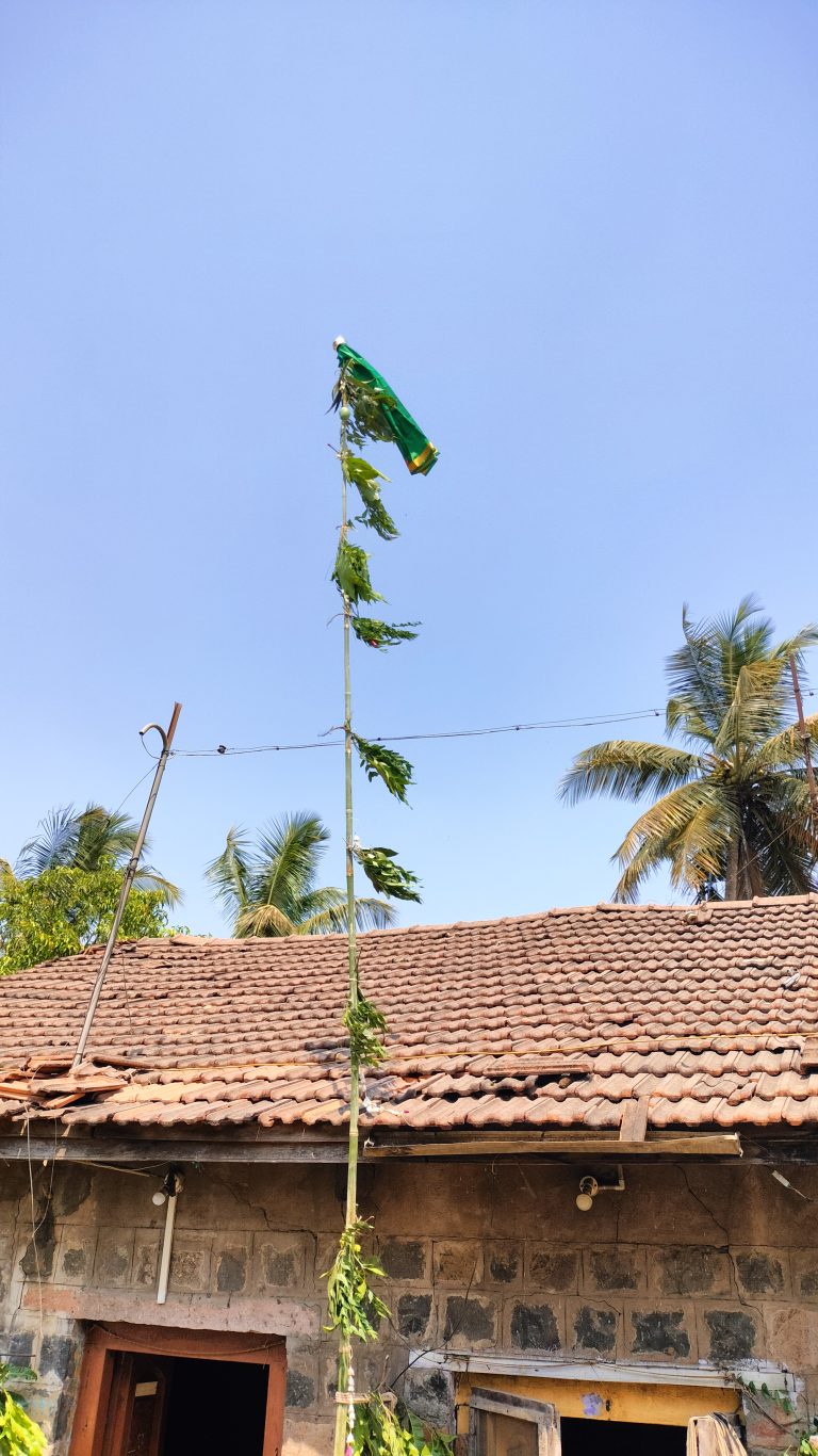 A weathered flag, mostly green with some yellow, fluttering at the top of a tall pole, with a backdrop of clear blue sky. The pole is in front of a traditional building with a red tiled roof and stone walls, with some green foliage and palm trees visible in the scene. (Gudi padwa festival)