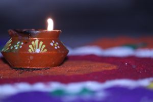 A lit terracotta diya with painted designs on a colorful rangoli background, reflecting the celebration of Diwali an Indian festival.