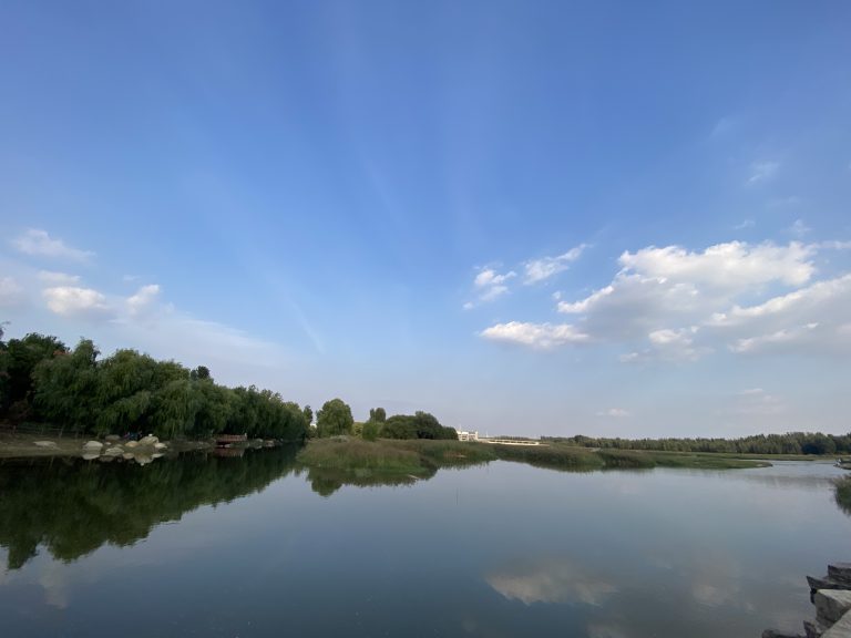 A serene landscape featuring a calm body of water reflecting the sky, surrounded by lush greenery under a blue sky with dispersed clouds and light rays emerging from the clouds.