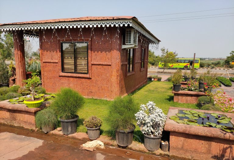 An attractive reception room greenhouse surrounded by a well-manicured garden featuring potted plants, a small pond with lily pads, and a lush green lawn. In the background, a road with a moving bus is visible.