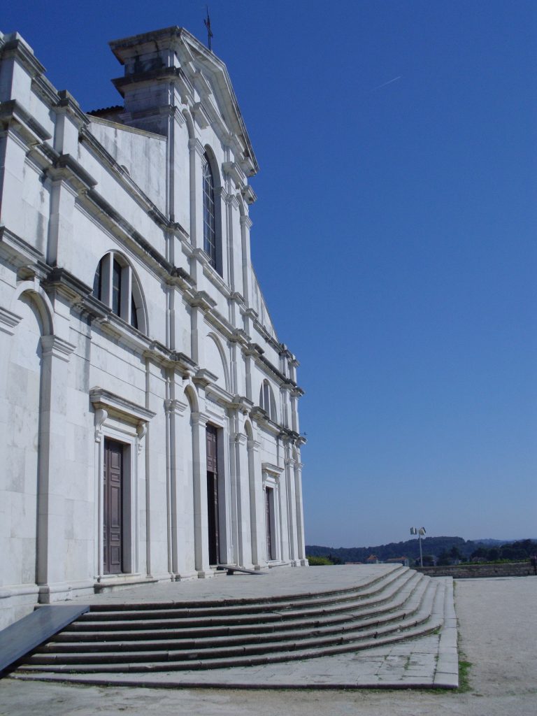 White baroque church facade with multiple columns and arched windows under a clear blue sky, fronted by a wide staircase and an open plaza.