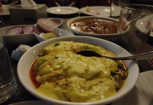 A close-up of a punjabi dish with a creamy green sauce, accompanied by other dishes including one with sliced onions and limes, a dark sauce, empty plates, a paper napkin holder, and a half-full glass of water on a dining table with dim lighting.