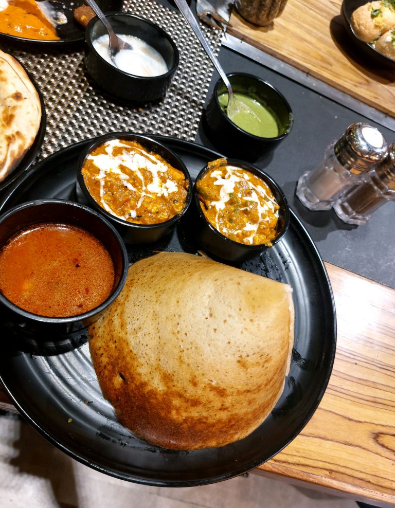 An overhead view of an Indian meal on a black plate, including a crispy dosa with various accompaniments such as coconut chutney, sambar, and other curry dishes, all arranged on a wooden table.