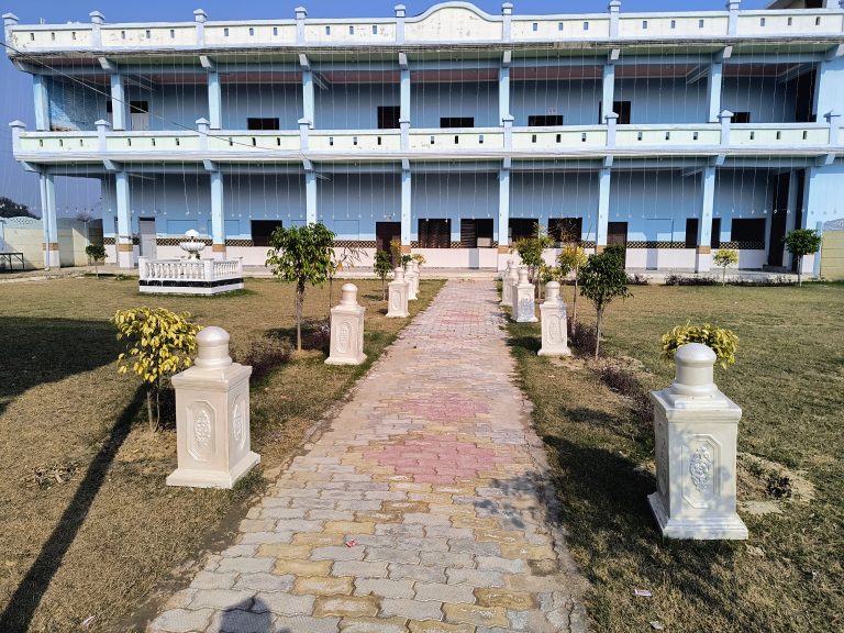 A paved pathway flanked by decorative white pillars leading towards large two-story building with a blue and white facade with greenery around.