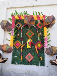A vibrant wall decoration featuring green foliage as the background, adorned with colorful flower garlands in shades of pink, orange, and yellow, and geometric patterned diamond-shaped hangings. Decorative umbrellas with intricate designs are mounted at the corners, adding to the festive look.