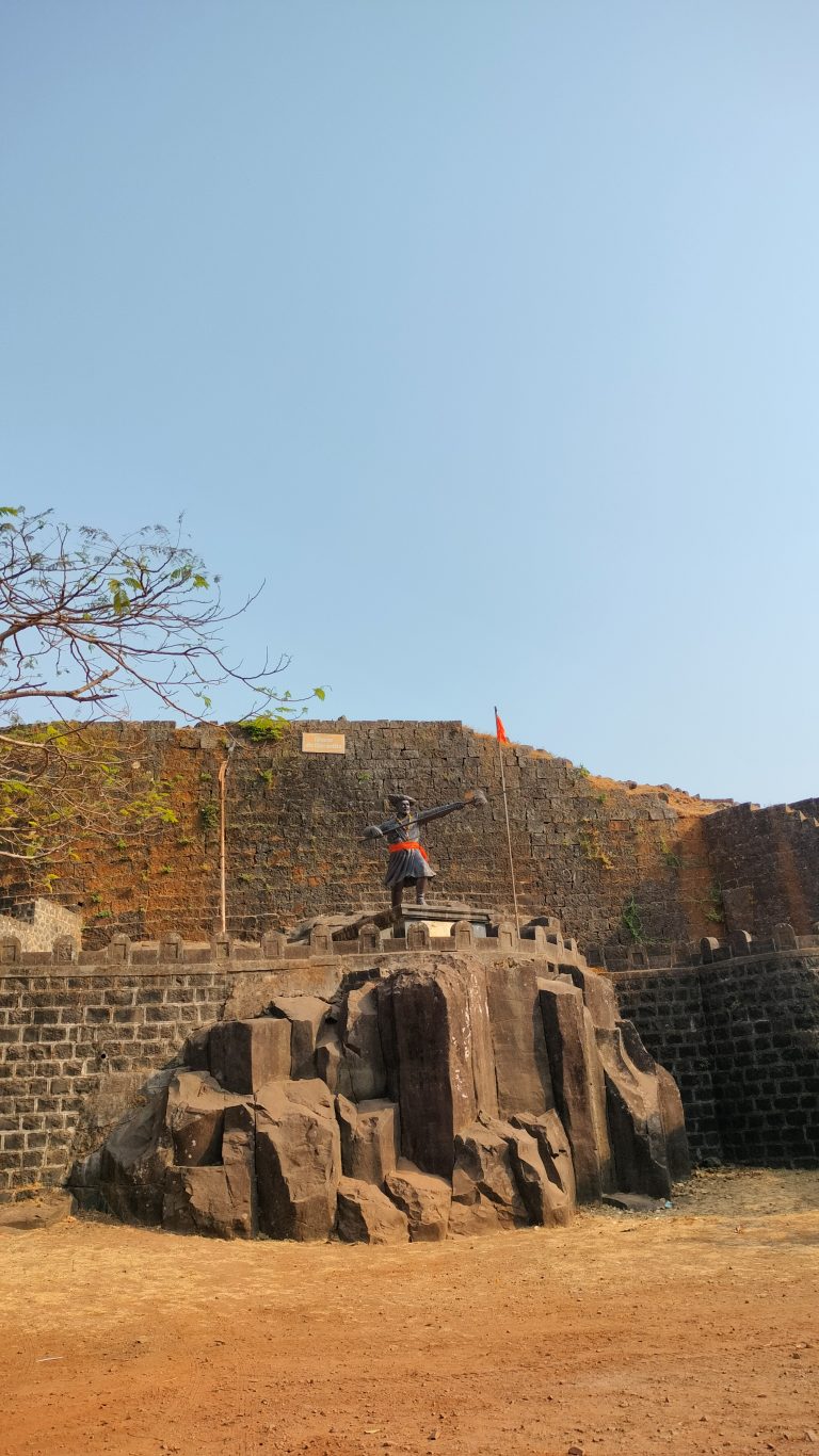 A large statue of Veer Shiva Kashid with a sword stands in front of an ancient fort with stone walls under a clear blue sky. Two orange flags are visible on top of the fort wall.