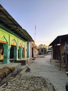 This image shows a serene village scene with traditional houses, paved pathways, and a flag hoisted on top of one of the buildings under a clear blue sky.