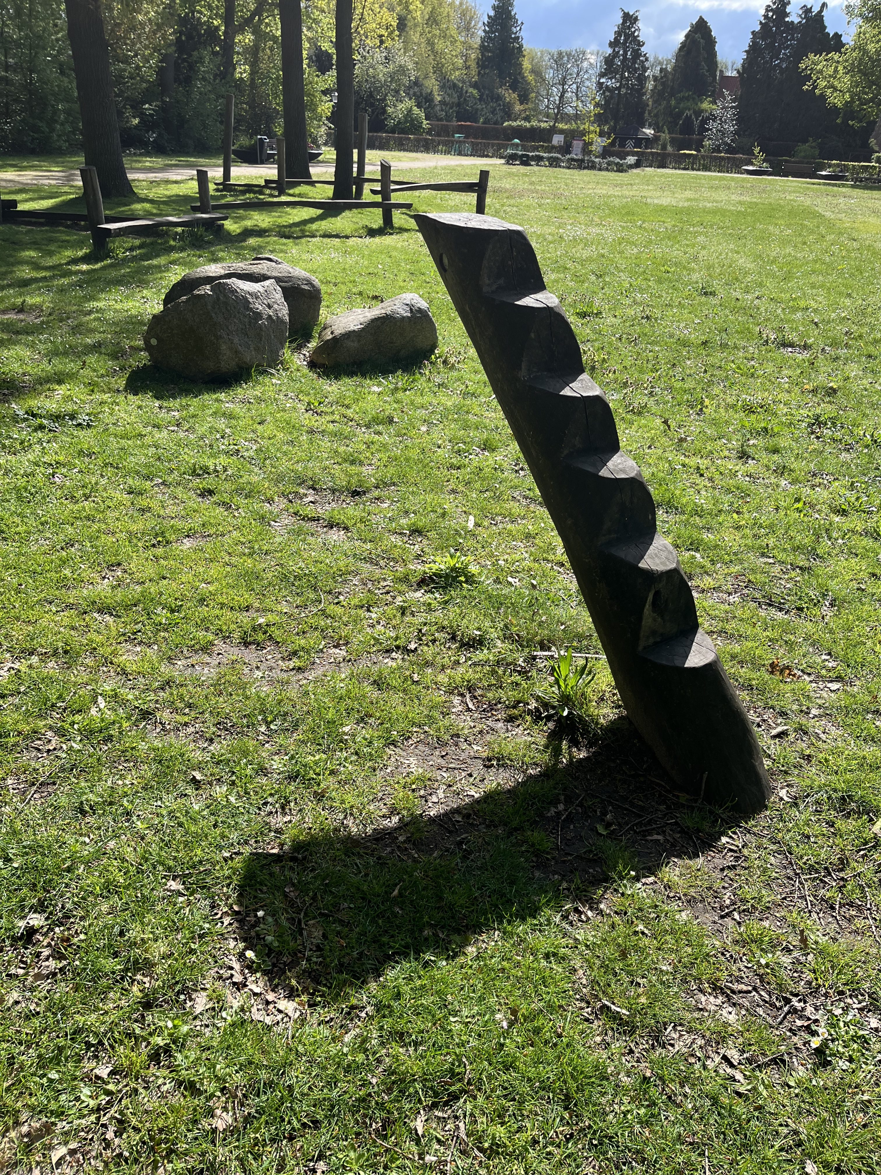 A Woden pole in the gras with steps on it and it’s shade on the ground