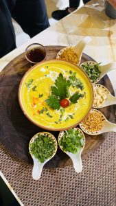 A colorful bowl of khao soi soup with a swirl of cream, garnished with herbs and a cherry tomato, surrounded by small dishes of various toppings and a cup of tea, served on a wooden platter on a woven table mat.
