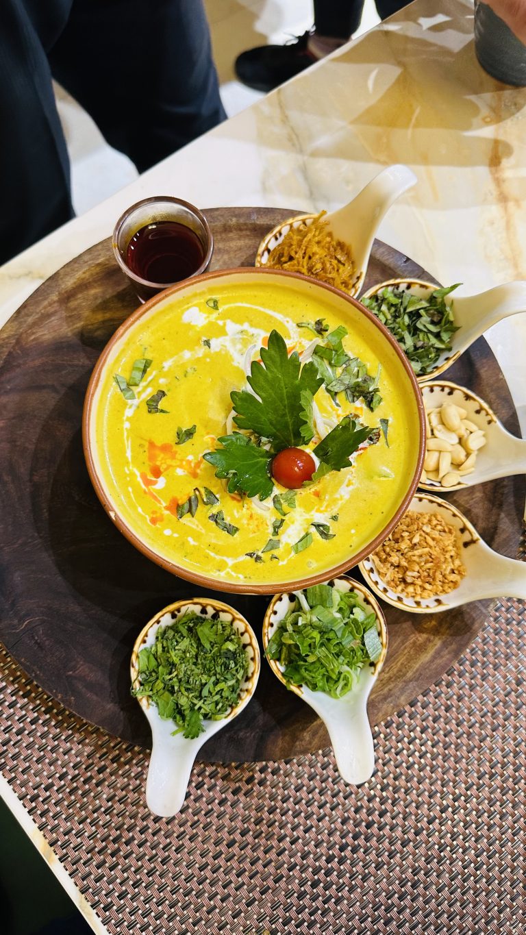 A colorful bowl of khao soi soup with a swirl of cream, garnished with herbs and a cherry tomato, surrounded by small dishes of various toppings and a cup of tea, served on a wooden platter on a woven table mat.