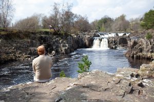 Man sat on the edge of the a cliff with a waterfall in the background 