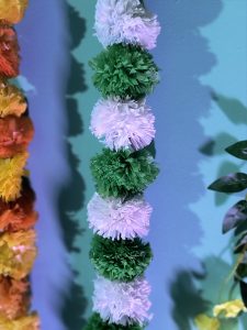 Artificial decorative pom-pom garland with colorful white, green, and orange pom-poms hanging against a turquoise wall, adding charm to the ambiance.