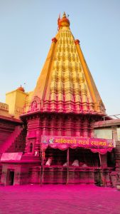 A towering Jyotiba temple with intricate carvings and red flags at the top against a clear blue sky, its lower part and the surrounding ground are covered with vibrant pink powder, and banners with Devanagari script hang above the entrance.