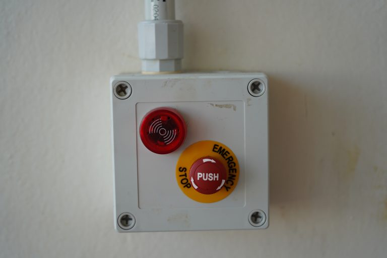 An emergency stop button installed on a white wall, featuring a red button with “EMERGENCY STOP” text and a yellow background. Above the button is a red indicator light with a concentric circle pattern.